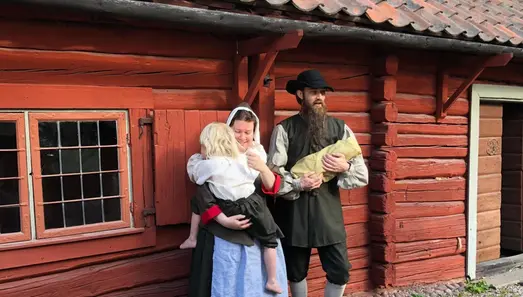 En familj i 1600-talskläder står utanför ett lågt rödmålat trähus. Mamman håller en pojke i famnen. Pojken är barfota. Pappan håller en bebis i famnen.